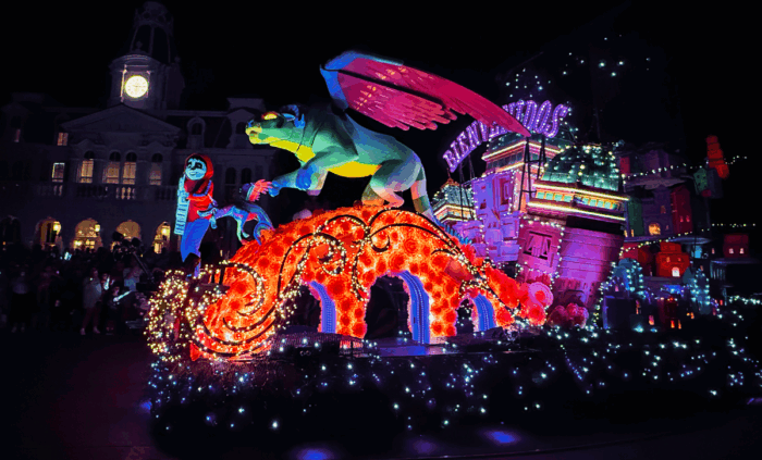 A bright red and purple parade float holds the characters Miguel, Pepita, and Dante from Disney's film Coco. The middle strurcture represents an arched bridge.